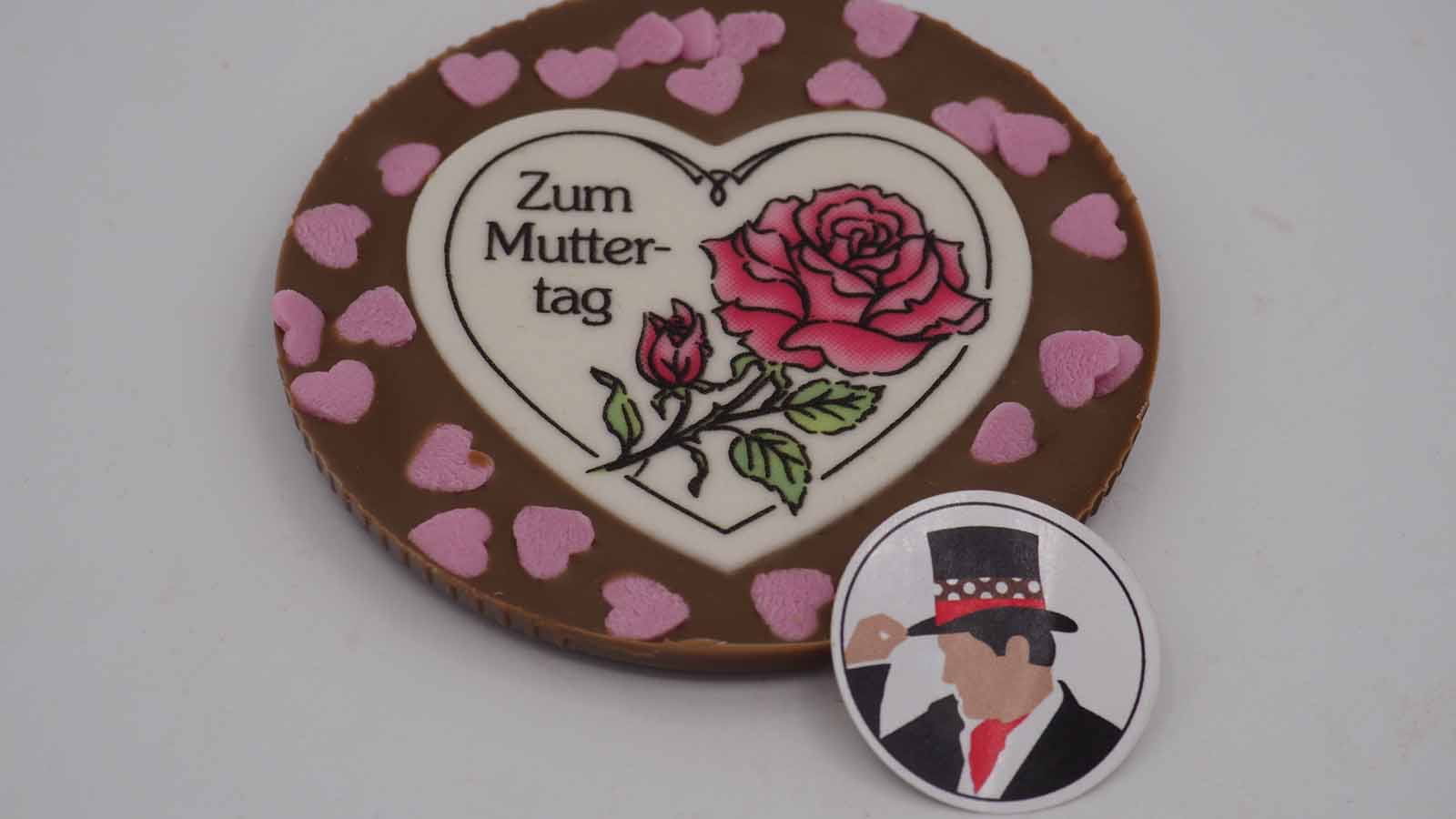 Handgezogene Scheibe aus Vollmilchschokolade mit Dekormasse (rosa Rose, Schriftzug: »Zum Muttertag«) und rosa Zuckerherzen zum Muttertag.